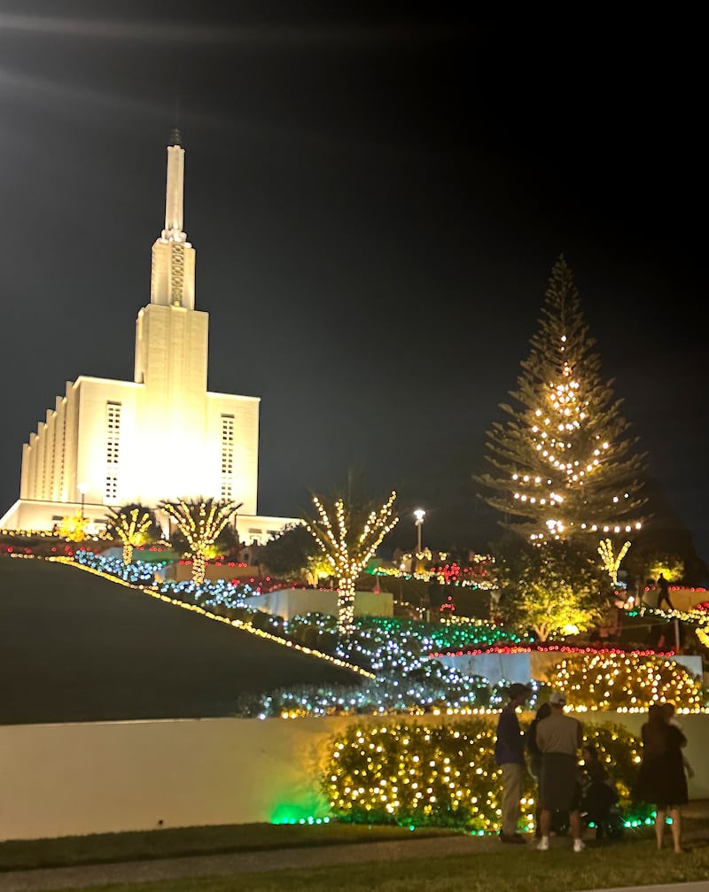 Os jardins do Templo de Hamilton Nova Zelândia estão iluminados com luzes de Natal nesta foto de dezembro de 2025. No primeiro plano, visitantes olham para uma cena da Natividade em frente ao templo.