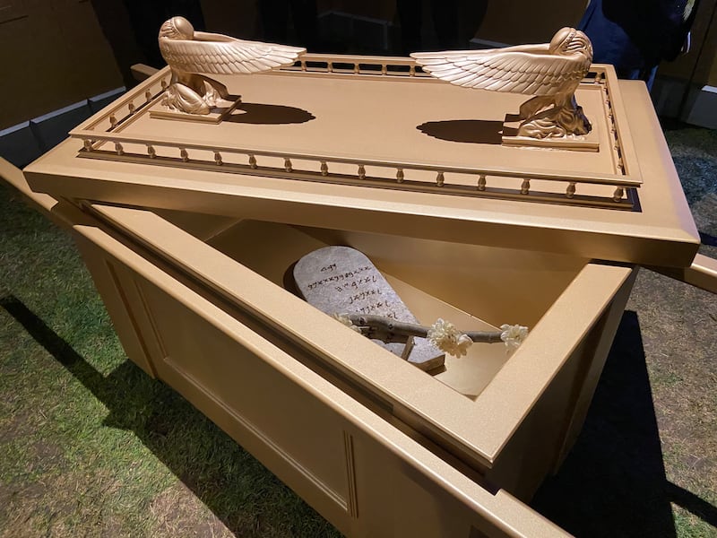 Uma réplica da arca dourada da aliança do tabernáculo antigo itinerante de Moisés, com duas estátuas de anjos no topo e com uma tábua de pedra e vara com flores dentro.