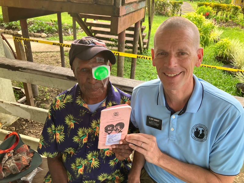 Pastor Ronny, à esquerda, tira uma foto com Élder Bret Dandoy, à direita, enquanto segura seu livreto de registro médico após cirurgia de catarata em 2026.