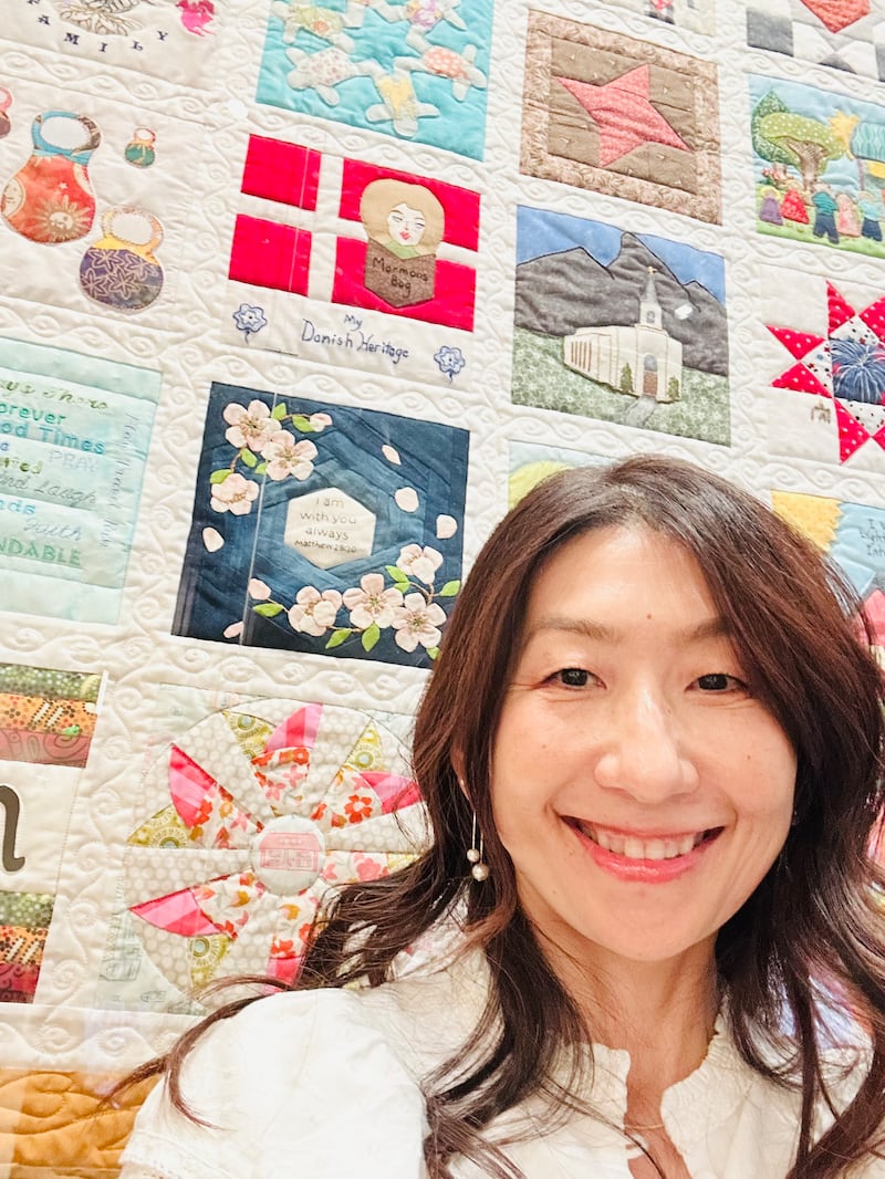 Sachiko Aldous fica ao lado do seu bloco de colcha, que é azul com flores de cerejeira aplicadas em cima, na exposição "The Family Quilt" no Centro de Visitantes da Praça do Templo em Salt Lake City, julho de 2026.