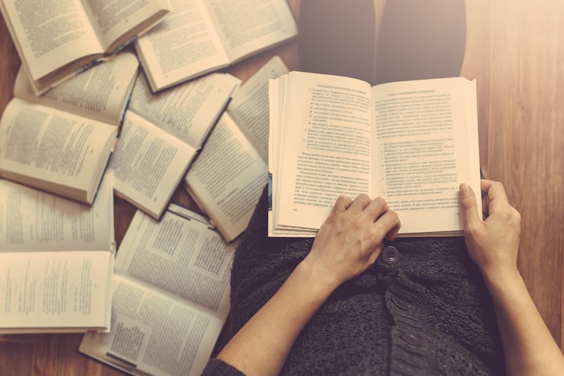 Mulher lê um livro com vários livros no chão ao redor dela.