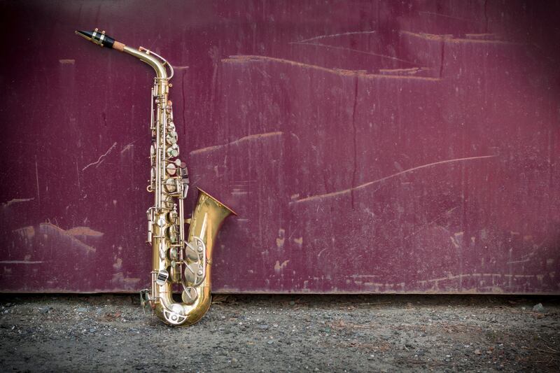 Saxofone alto apoiado no chão, encostado em uma parede vermelha.