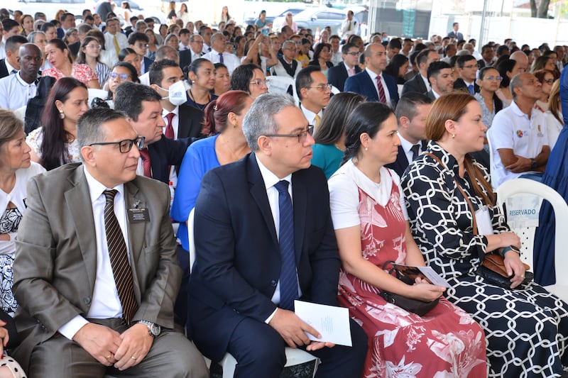Uma grande congregação de pessoas sentadas em cadeiras ao ar livre em trajes de domingo.