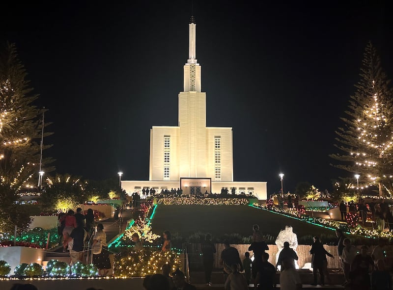Os terrenos do Templo de Hamilton Nova Zelândia estão iluminados com luzes de Natal nesta foto de dezembro de 2025. No primeiro plano, os visitantes observam uma cena de Natividade em frente ao templo.