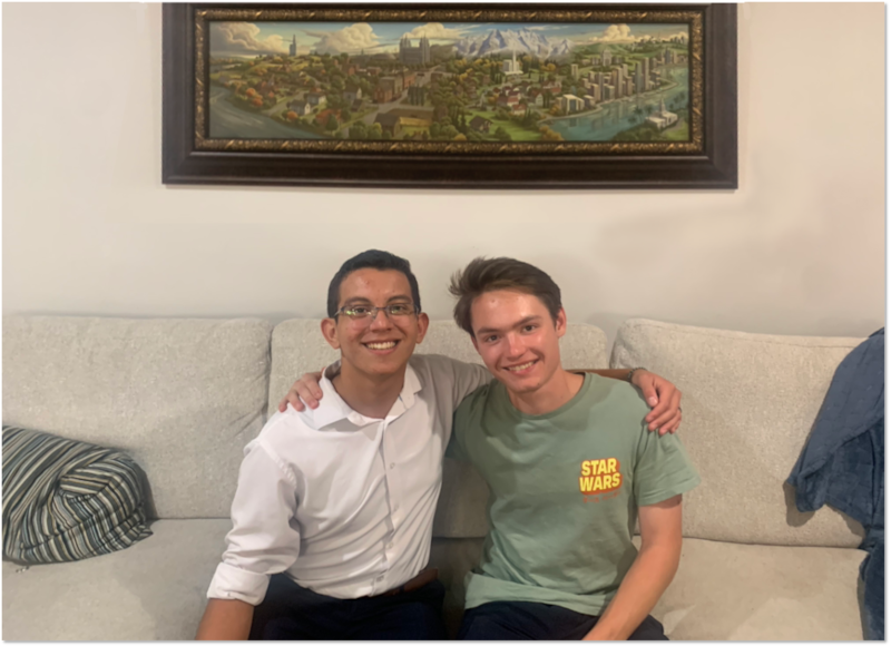 Oscar Hernandez e Benjamin Woodworth tiram uma foto juntos na casa dos Woodworth em Utah com a capa de "Saints" na parede atrás deles em 2024.