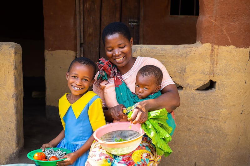 Uma menina senta-se com a mãe, que carrega seu irmão de 1 ano enquanto corta legumes fora de uma casa no sul do Malawi.