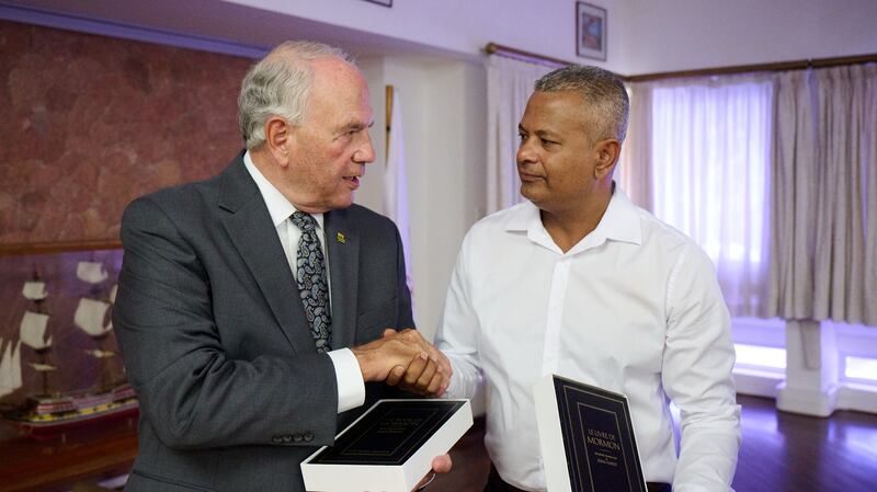 Elder Ronald A. Rasband aperta a mão de Sebastien Pillay, o vice-presidente das Seychelles, enquanto seguram cópias do Livro de Mórmon em 29 de outubro de 2025.
